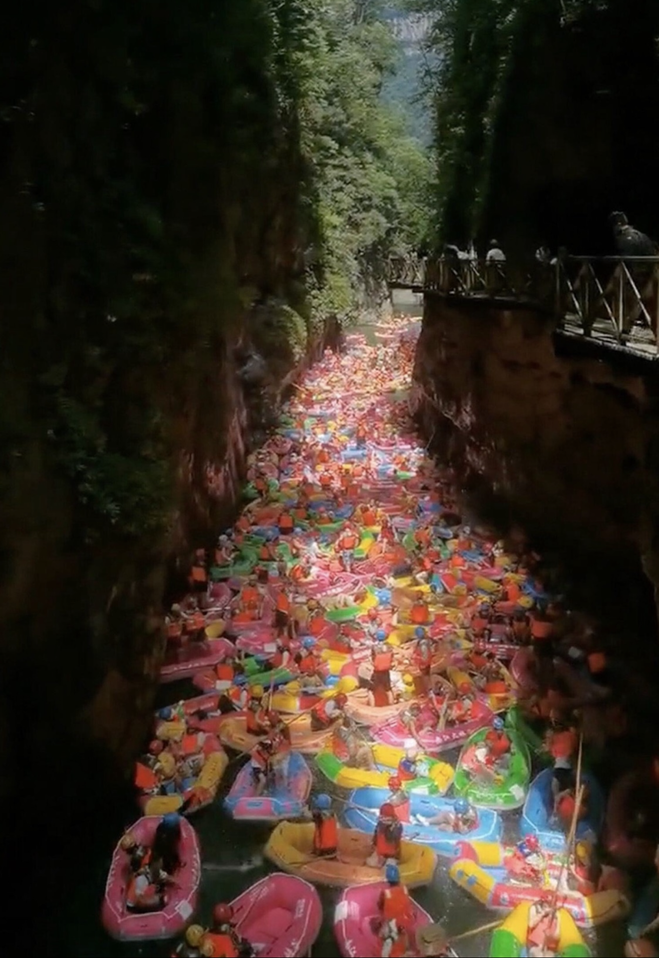 A narrow canyon with high rocky walls and a wooden walkway on the right side, filled with numerous colorful inflatable boats and people swimming in the water below.