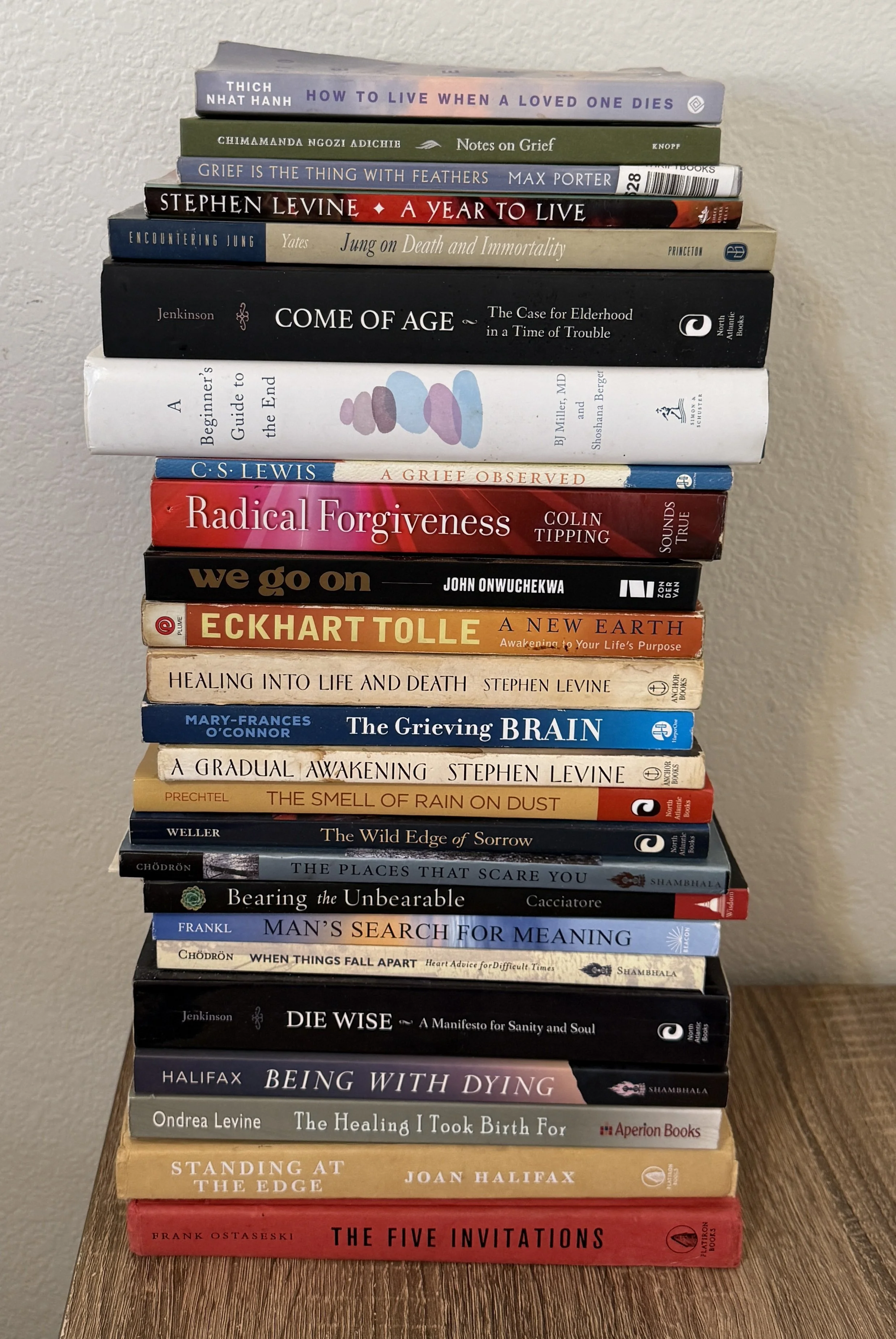 Stack of multiple books on a wooden surface against a wall, featuring titles related to grief, life, death, and personal growth.