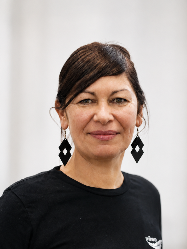 A woman with short brown hair wearing a black shirt and large black geometric earrings, standing against a plain light background.