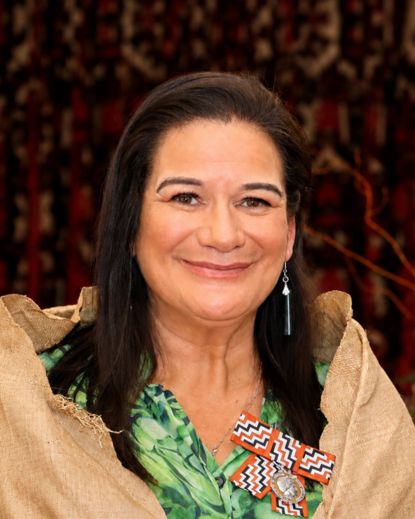 A woman with long dark hair smiling, wearing a green patterned shirt, patterned ribbon with a 'Dream' medal, and a beige outer garment, with a colorful background.