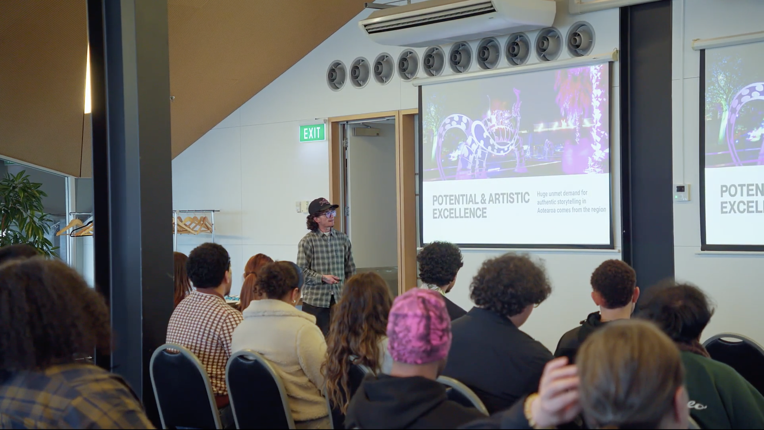 A man in a plaid shirt and baseball cap giving a presentation in a conference room filled with attendees. A large screen displays a slide titled 'Potential & Artistic Excellence' with a colorful background, and there is a smaller screen showing the same content. The room has an exit sign above the door and air vents on the ceiling.