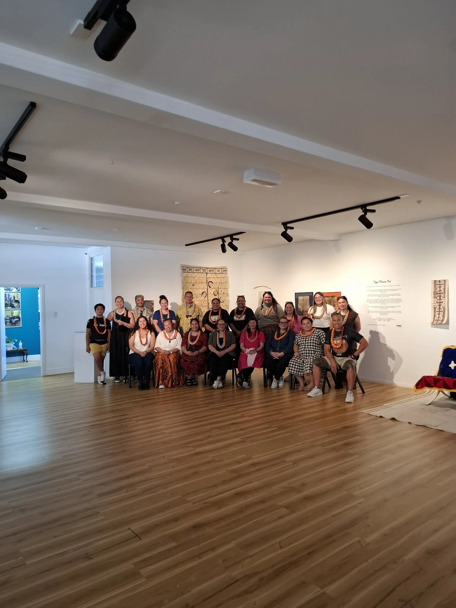 A group of diverse people standing and sitting in an art gallery, wearing traditional clothing and jewelry, with some smiling at the camera.
