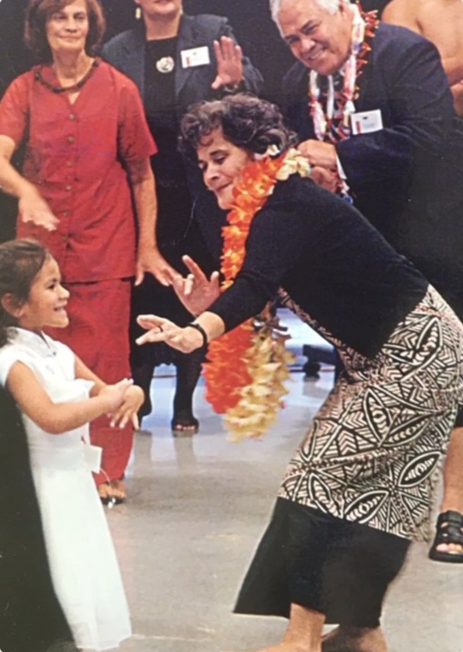 A woman with short curly hair, wearing a black top, patterned skirt, and orange lei, is seemingly dancing or playing with a smiling young girl in a white dress at a social gathering. Several adults are standing around, including a woman in red and a woman with gray hair in a dark jacket with leis, in what appears to be an indoor celebration or party.