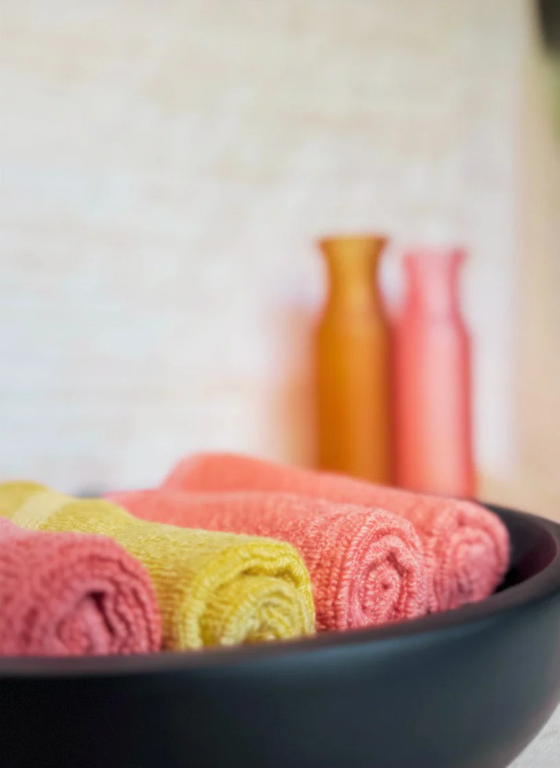 Colorful rolled towels in a black container with blurred orange and pink vases in the background.