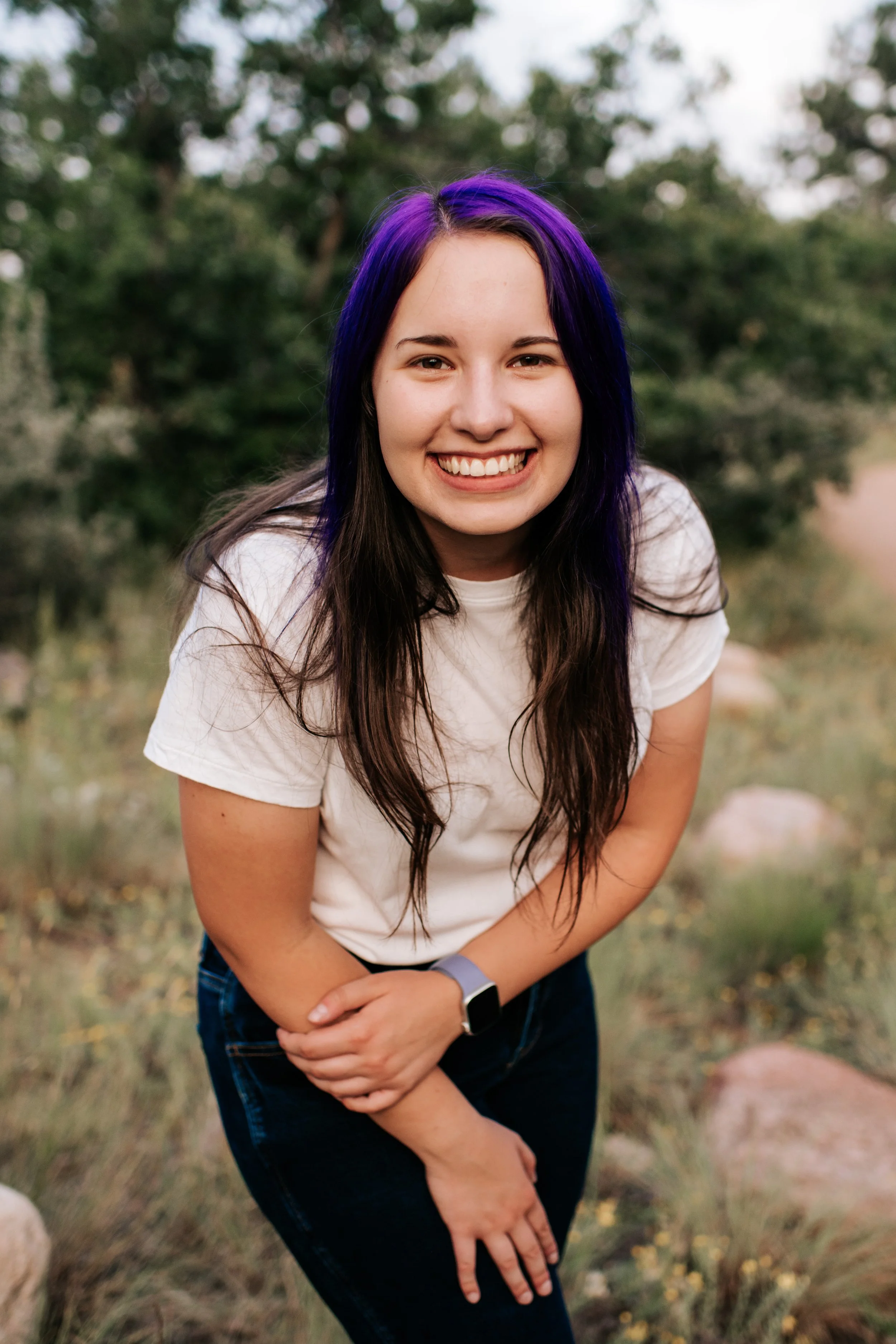 A young woman with long dark hair and purple highlights, smiling and leaning forward outdoors during daytime, with trees and grass in the background.