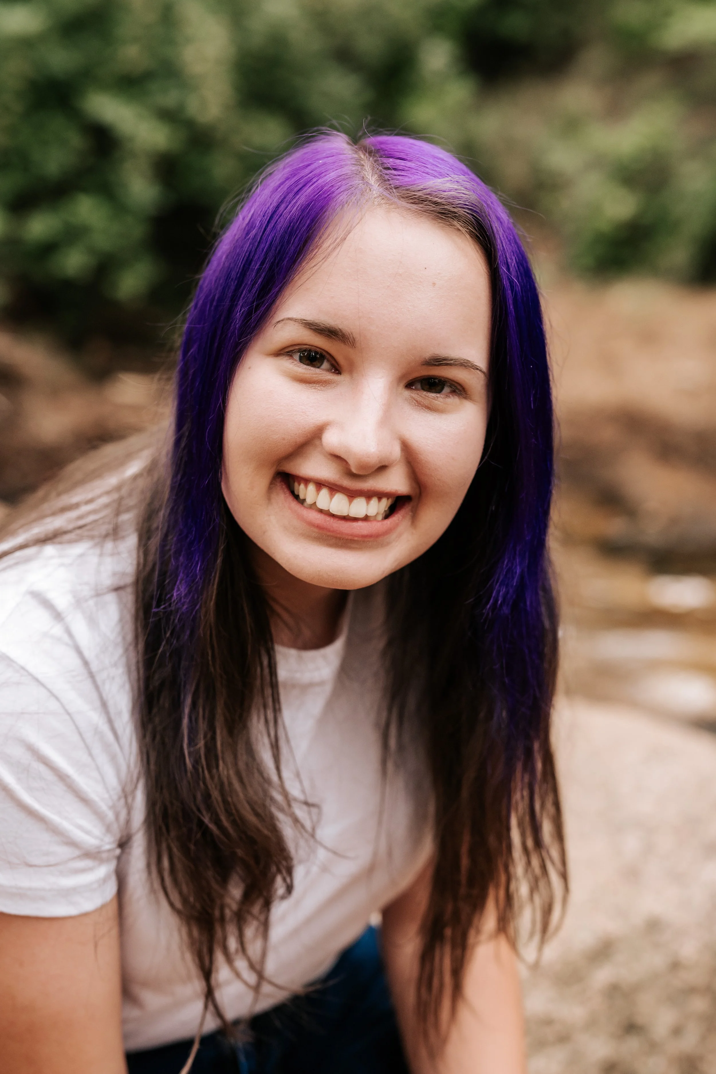 Close-up of a young woman with purple and black hair, smiling outdoors.