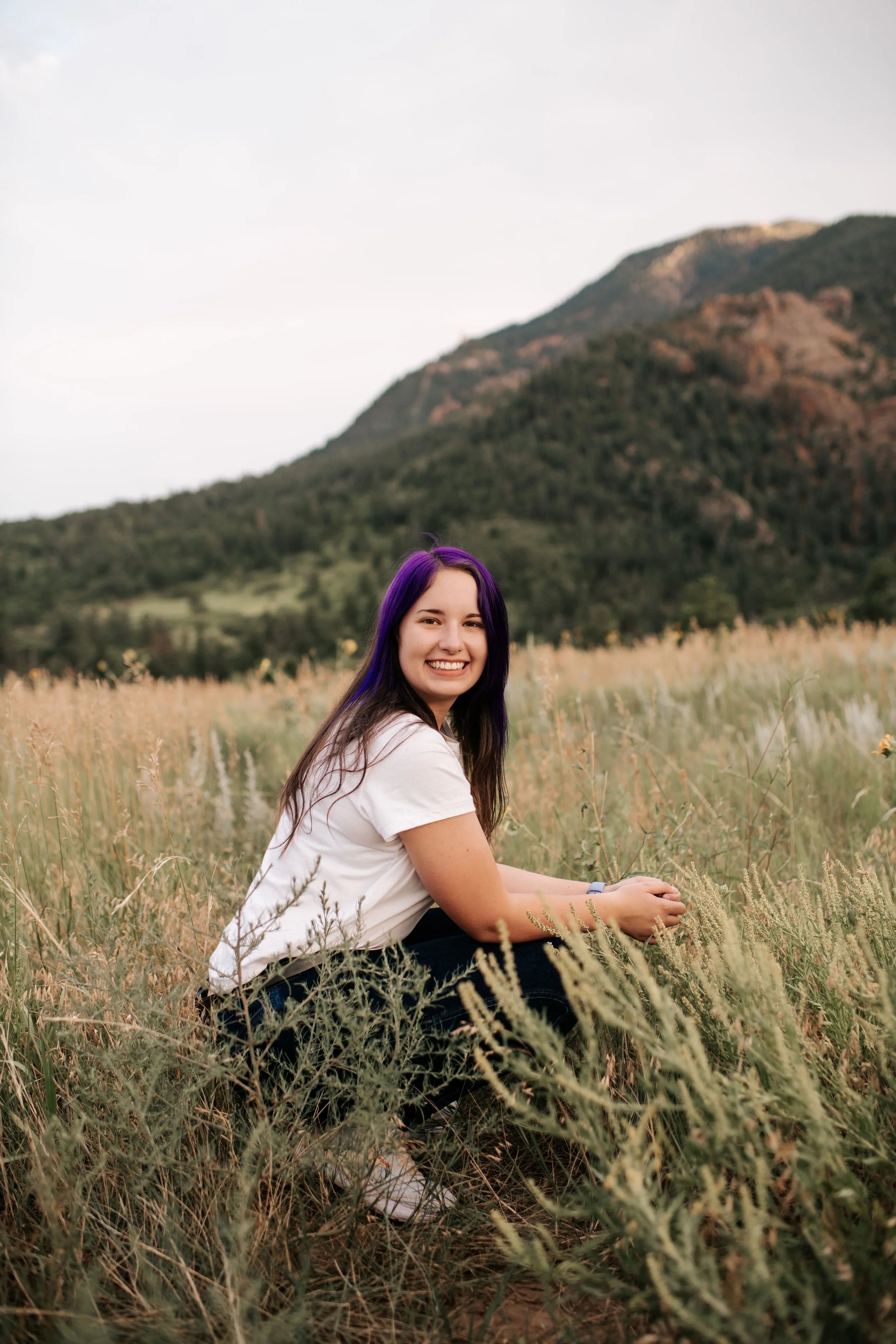 A young woman with purple hair sitting in a field of grass with mountains in the background, smiling at the camera.