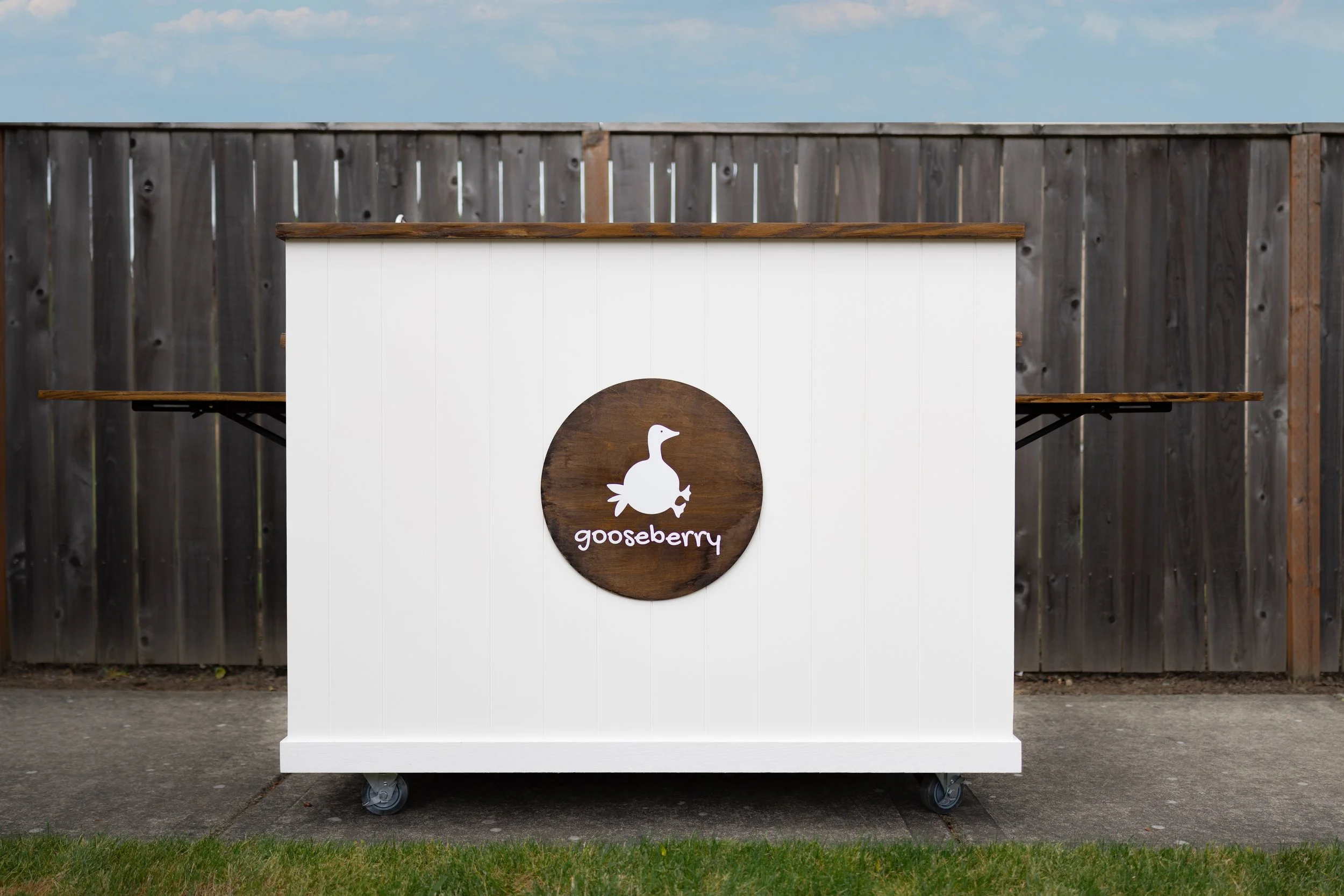 A white service cart with a circular sign featuring a duck silhouette and the word 'gooseberry' on it, set against a wooden fence and a partly cloudy sky.