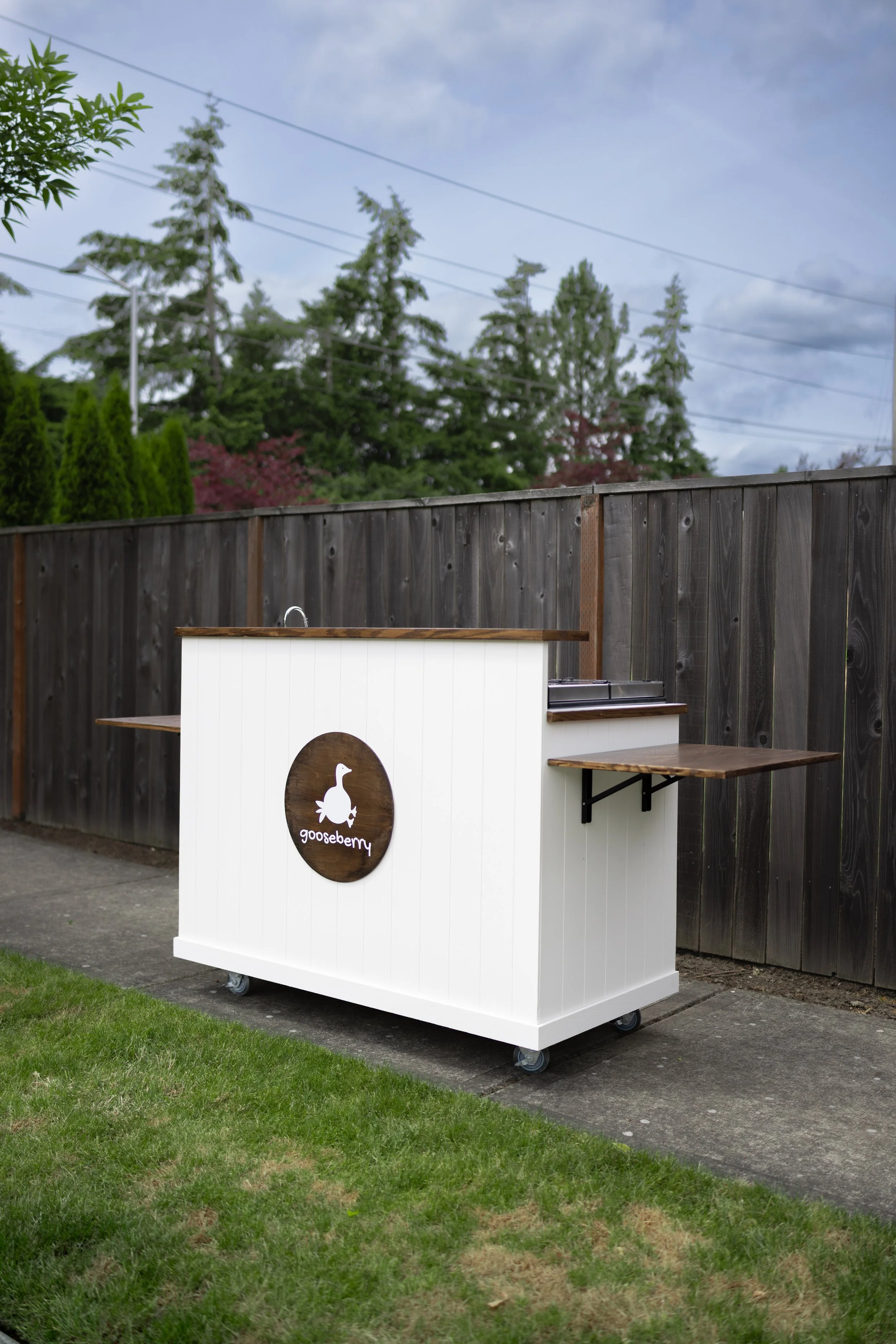 A white mobile cart with a gooseberry logo on it, situated outdoors on a paved surface near a grassy area and a wooden fence, with trees and power lines in the background.