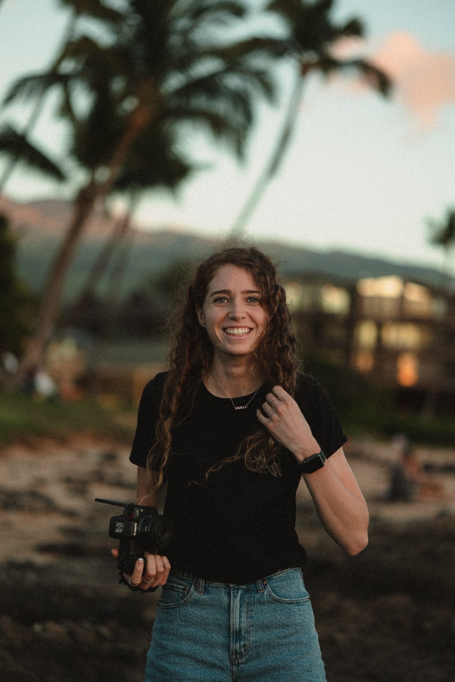 A young woman with curly brown hair smiling outdoors during sunset, wearing a black t-shirt, denim jeans, a watch, a necklace, and holding a camera stabilizer.