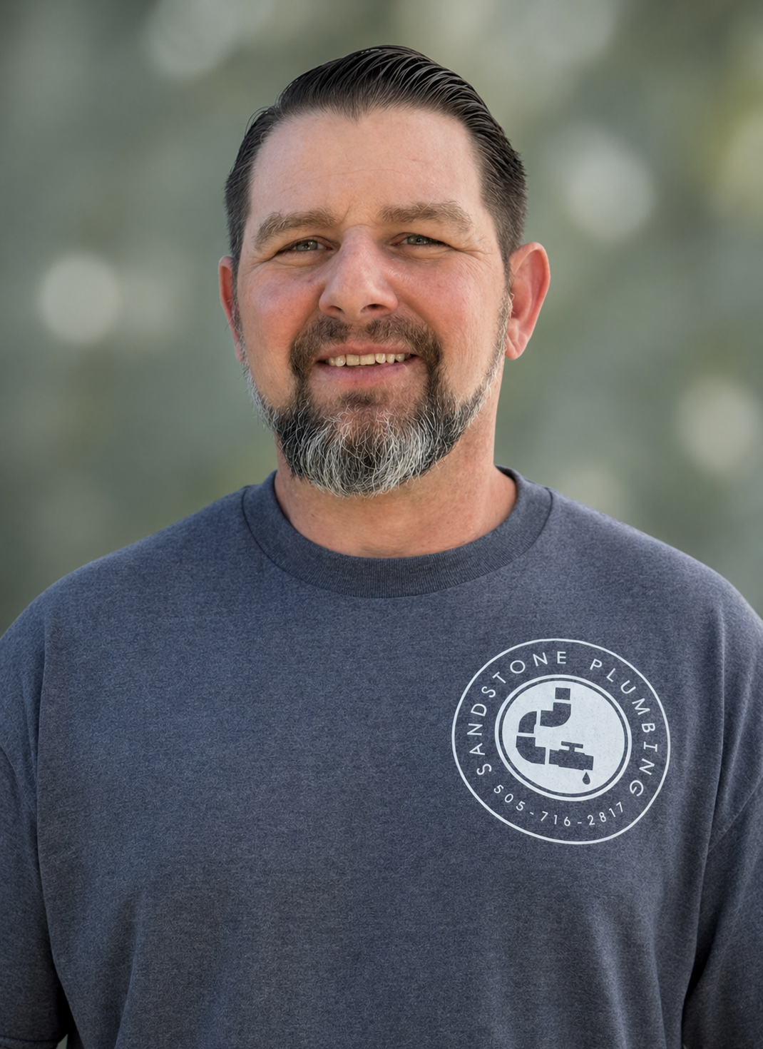 A smiling man with dark hair and a beard, wearing a gray sweatshirt with a logo that reads 'Sandstone Plumbing' and includes a faucet graphic, standing outdoors with a blurred background.