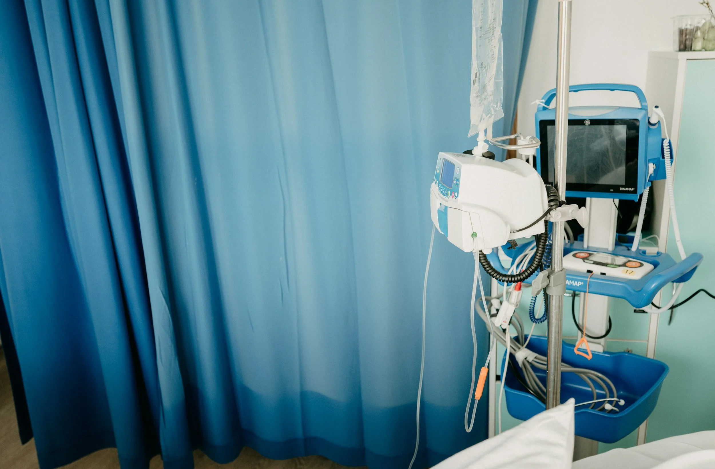 Hospital room with blue privacy curtain and medical monitoring equipment including a vital signs monitor and IV pole.