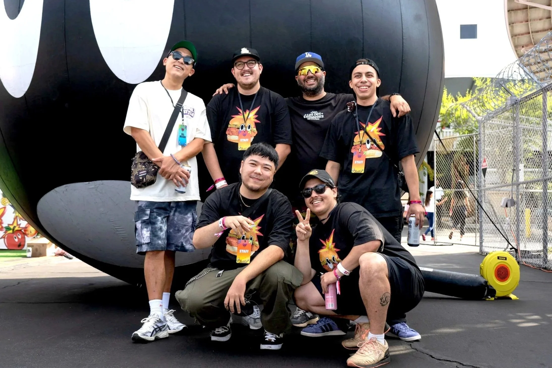 Group of five diverse young men posing and smiling in front of a large black and white panda sculpture at an outdoor event. Some are wearing sunglasses, staff lanyards, and casual summer clothing.