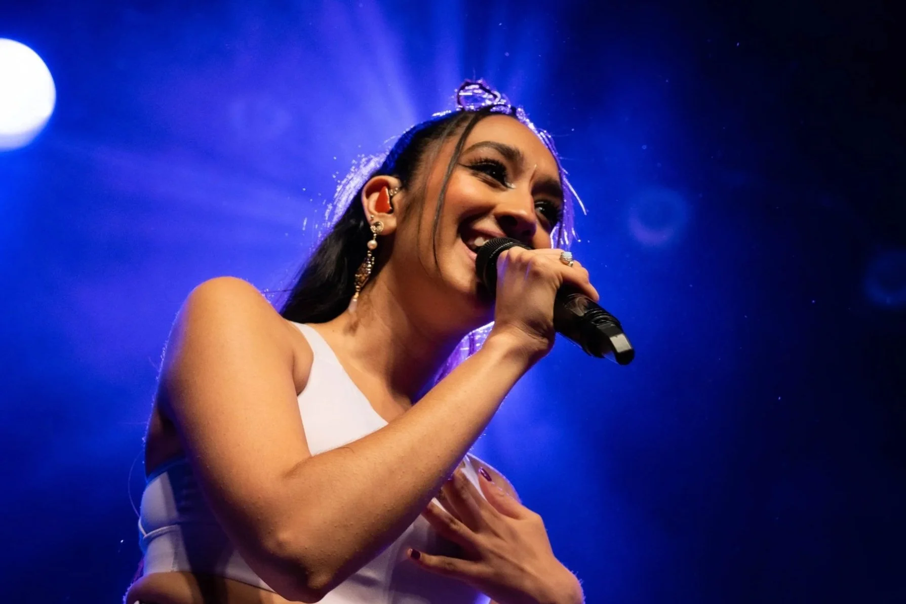 A woman with dark hair, wearing jewelry and a white top, is smiling as she stands near a microphone, with a colorful flower arrangement in front of her, and bright blue stage lighting in the background.