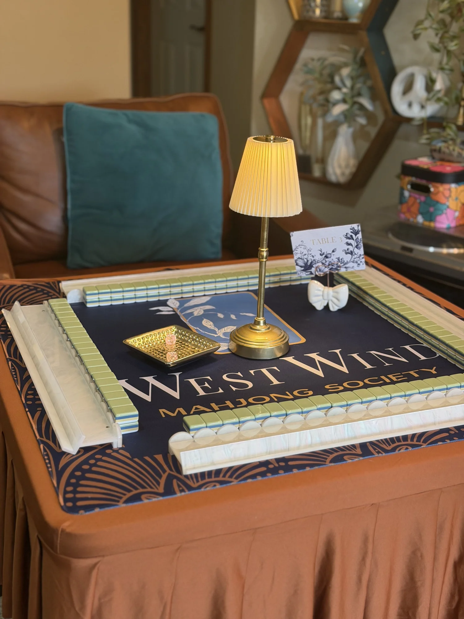 A West Wind Mahjong Society table set with racked tiles, dice, and a table number card