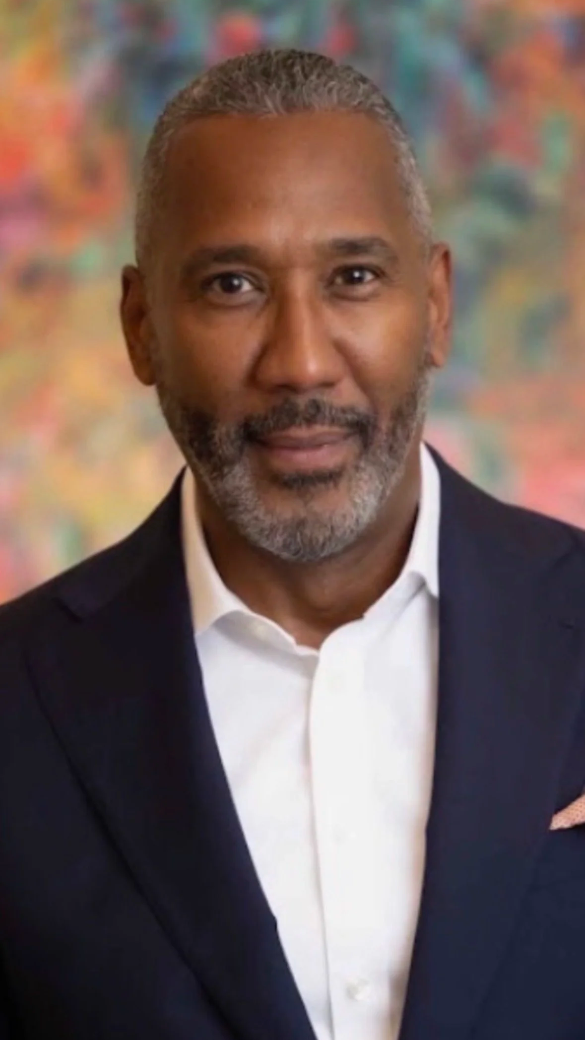 A portrait of a middle-aged man with short, gray hair and a beard, wearing a black suit jacket and white dress shirt, against a colorful, blurred background.