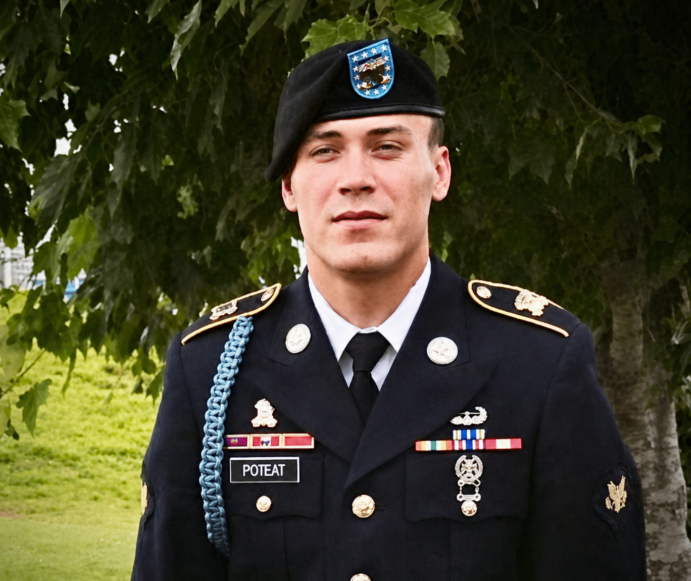 A young man in a military uniform standing outdoors with green trees in the background.