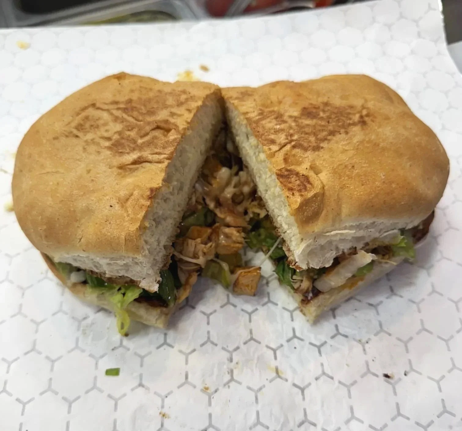 A torta cut in half, containing shredded chicken, lettuce, and vegetables inside a sandwich bun on a white hexagonal patterned surface.