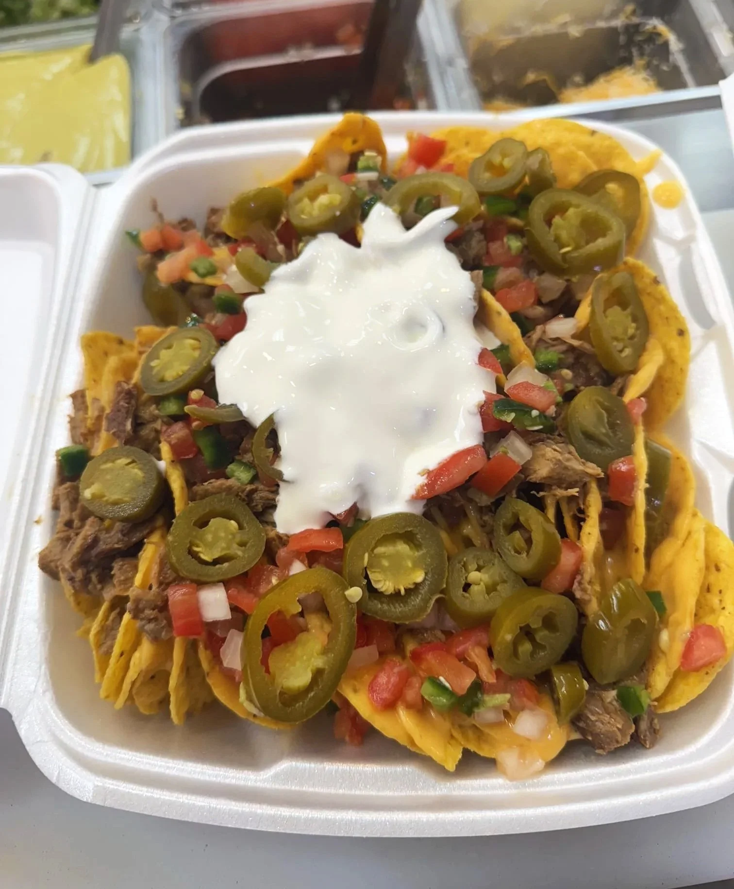 Close-up of a styrofoam tray of nachos topped with jalapeño peppers, diced tomatoes, onions, and sour cream.
