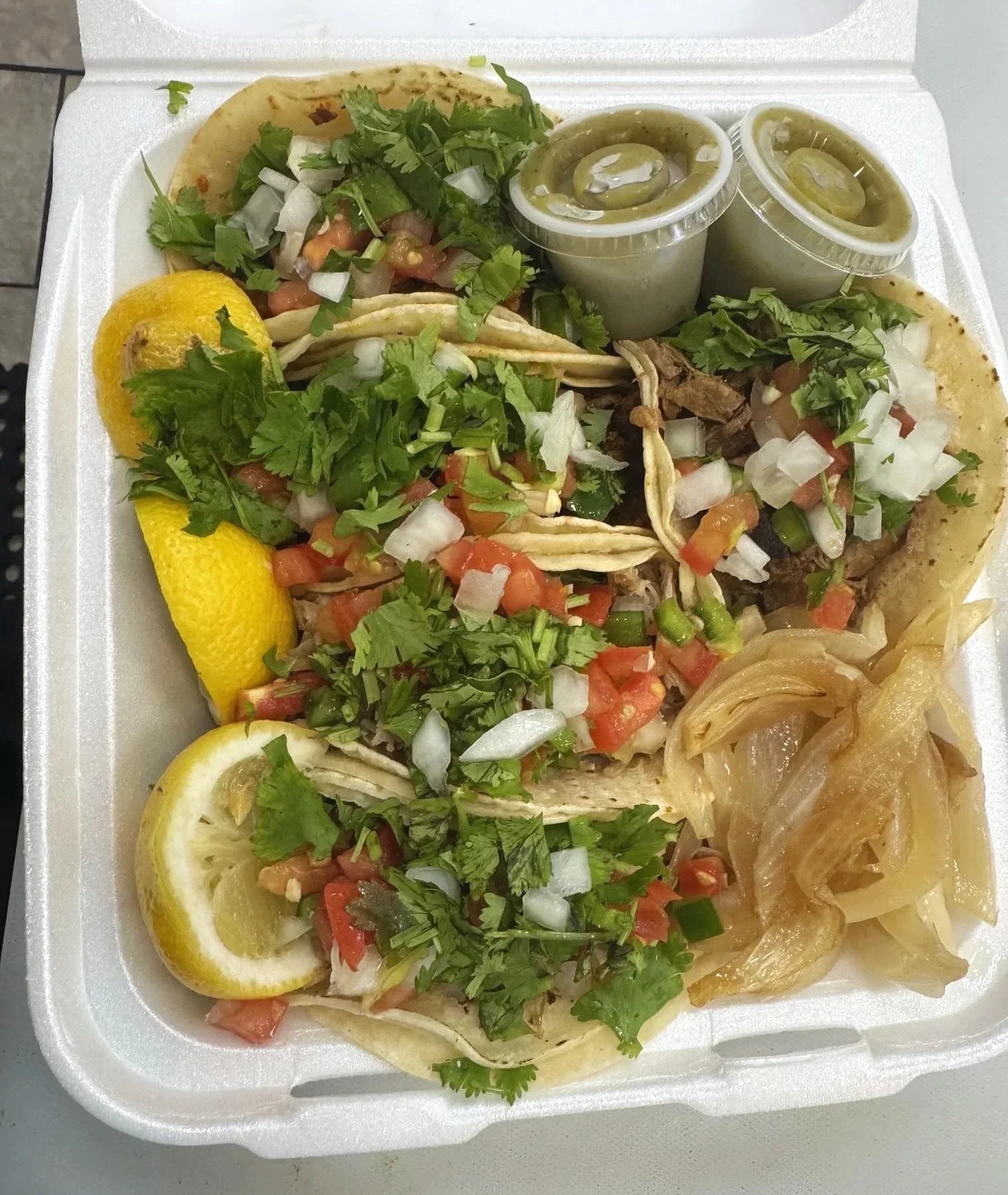 Five tacos filled with diced tomatoes, onions, cilantro, and meat, served with lemon wedges and sides of green sauce and caramelized onions, all in a styrofoam container.