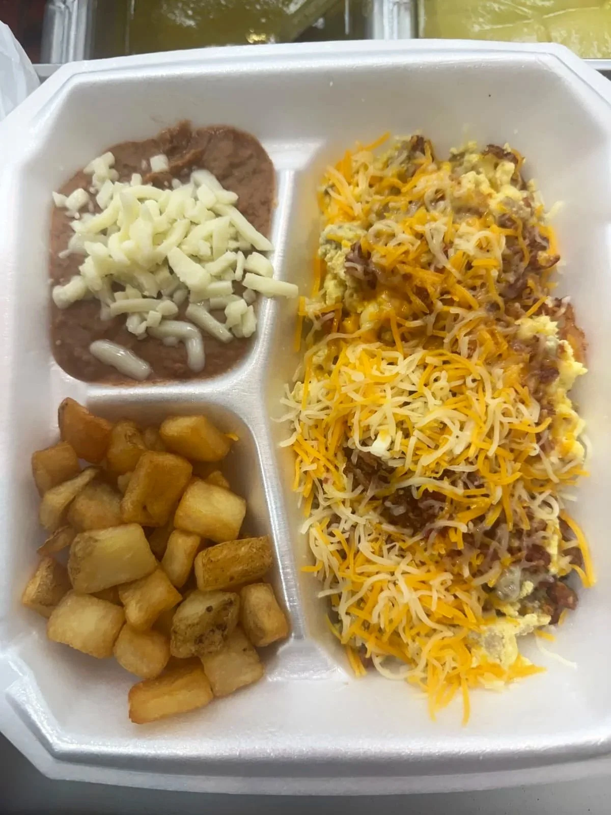 A foam tray with three compartments holding breakfast food including breakfast potatoes, meat with cheese and beef, and refried beans with cheese on top.