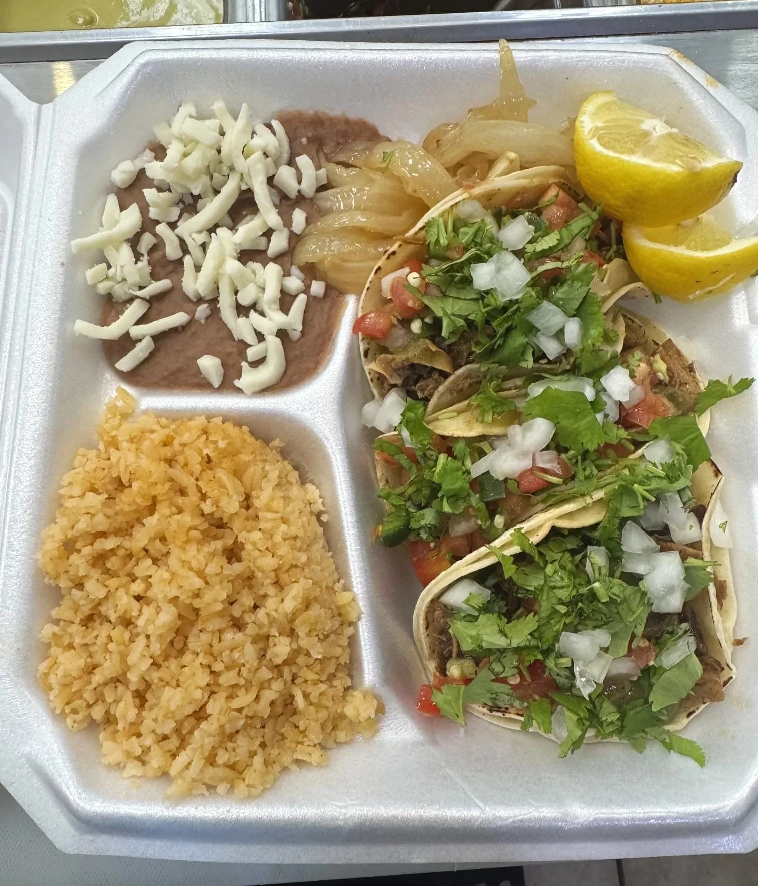 A styrofoam tray containing Mexican food with rice, refried beans topped with cheese, and three tacos filled with meat and topped with chopped onions, cilantro, and salsa, with lemon wedges.