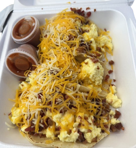 Breakfast tacos topped with shredded cheese, scrambled eggs, and bacon, with two containers of salsa on the side, served in a foam takeout container.