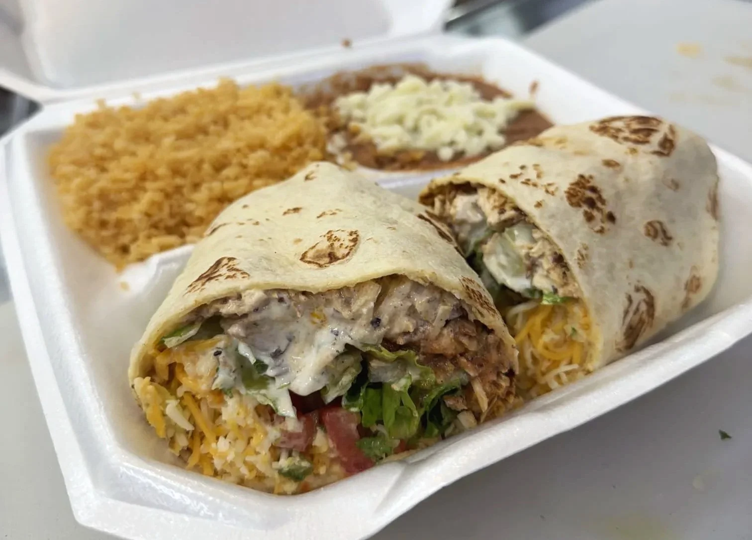 A takeout container with a burrito, rice, beans, and shredded lettuce and cheese.