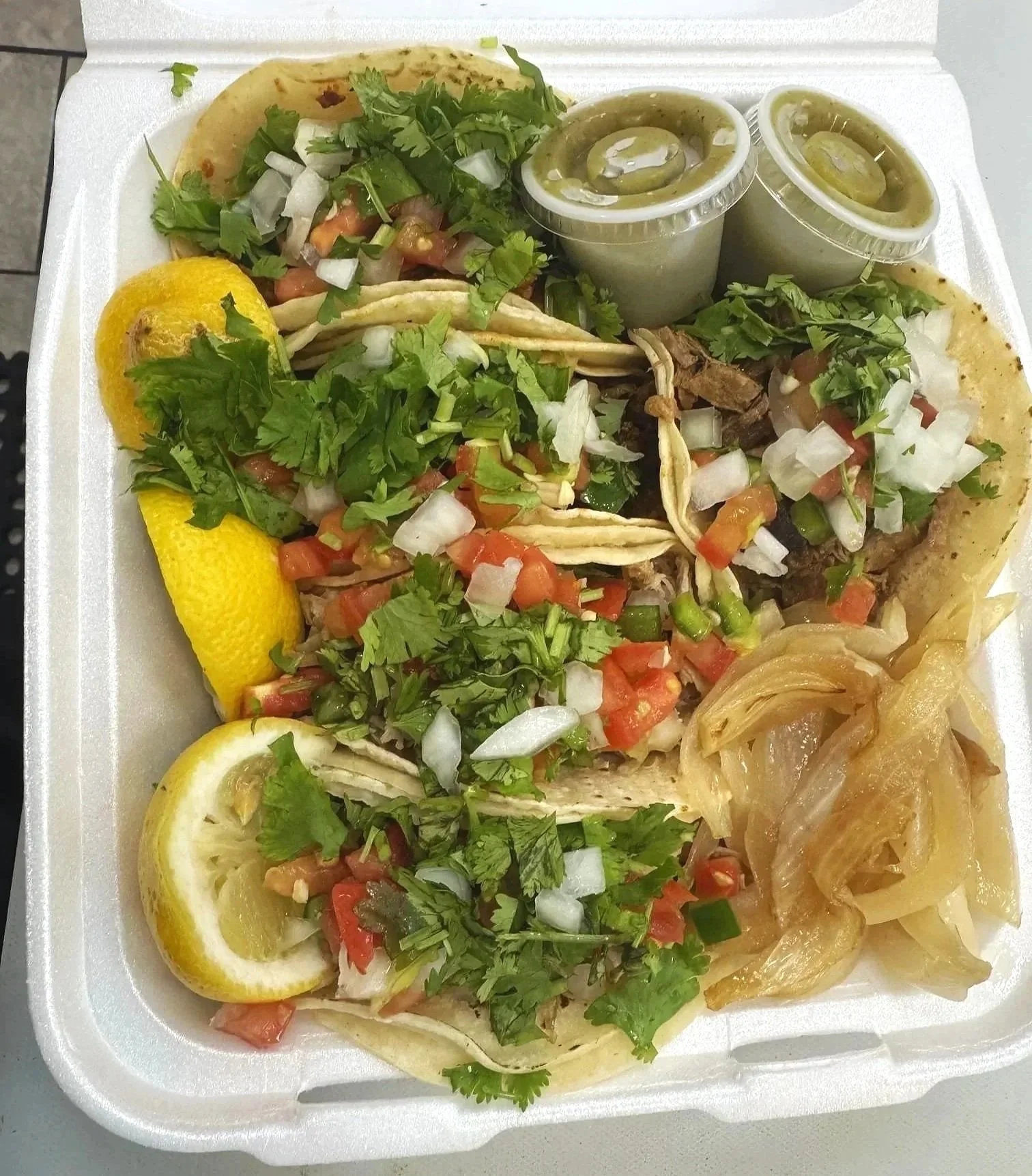 A styrofoam takeout container with five tacos filled with chopped onions, cilantro, diced tomatoes, and meat, accompanied by lemon wedges, green sauce in small containers, and caramelized onions.