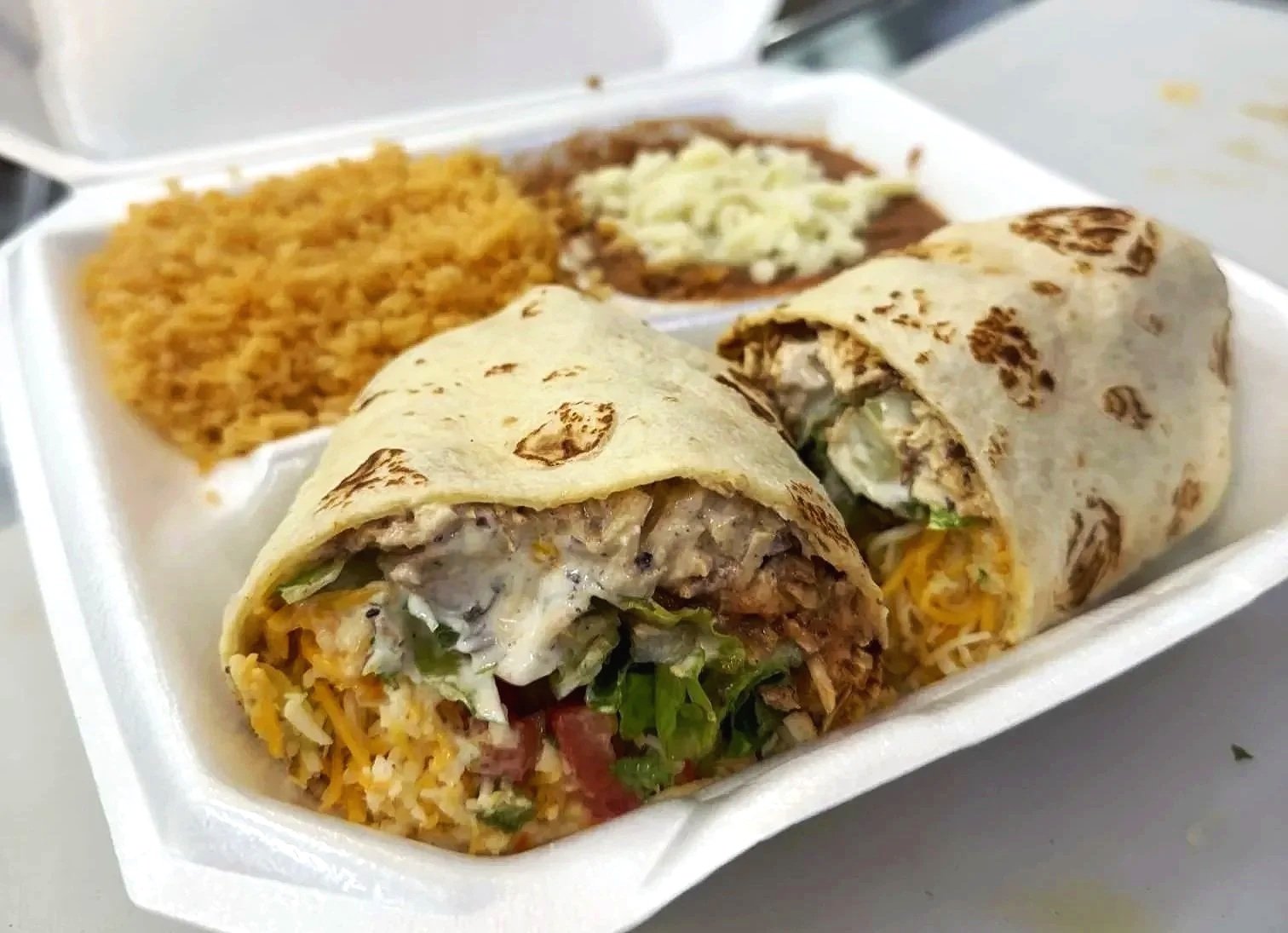 Burrito with shredded cheese, lettuce, and tomato, served with Mexican rice, refried beans topped with shredded cheese, and shredded lettuce with sour cream in a takeout container.