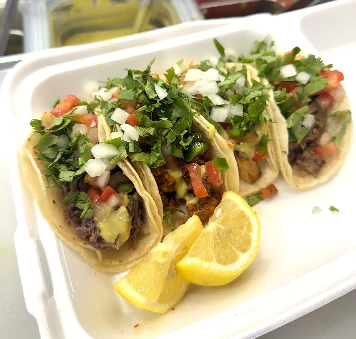 Four tacos filled with meat, topped with chopped onions, cilantro, and melted cheese, served with lemon wedges.