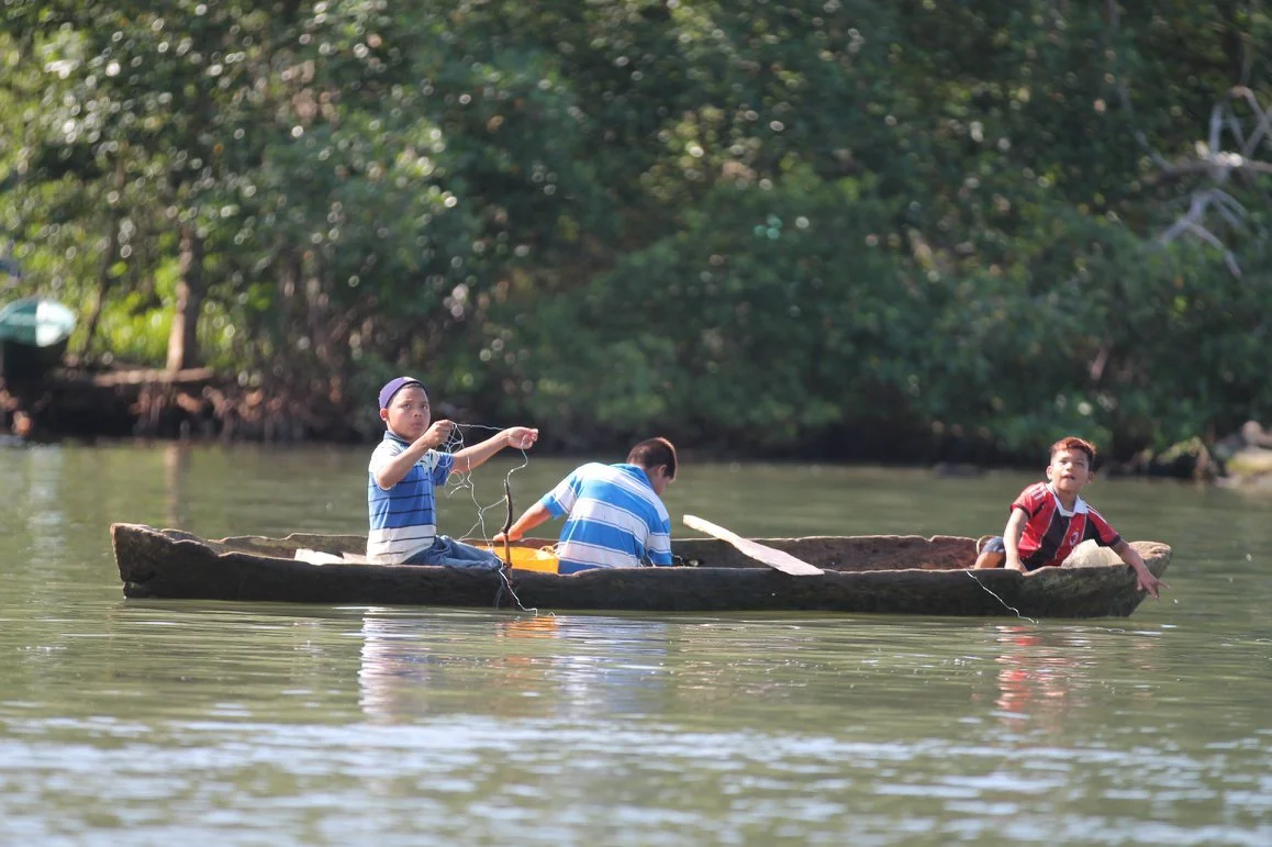 Three boys in a small wooden boat on a river, with dense green trees in background.