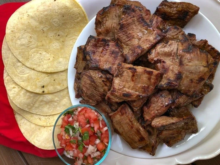 Grilled beef short ribs, corn tortillas, and a small bowl of chopped tomato and onion salsa on a red cloth.