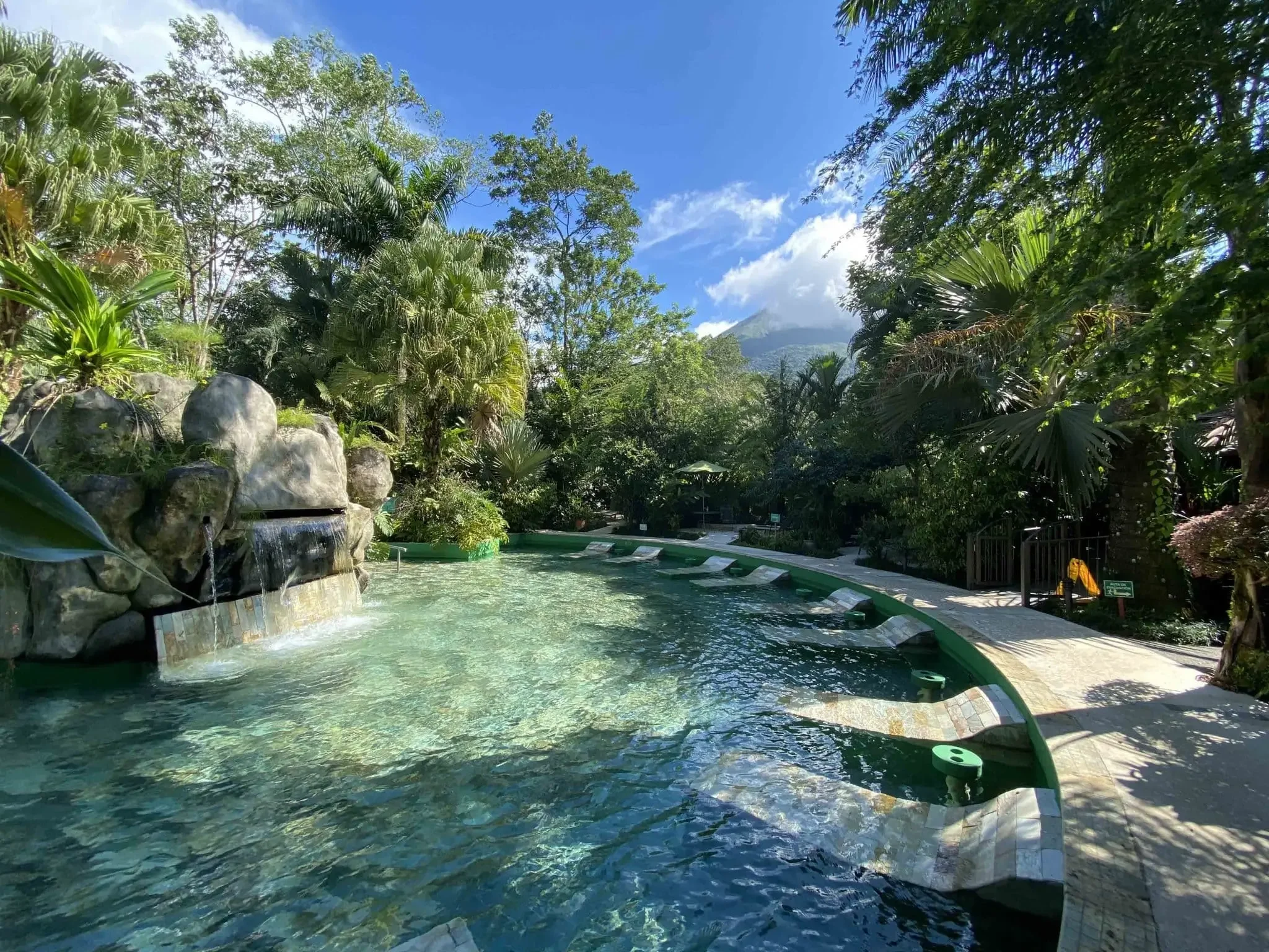 A tropical scene with a water feature, lush green trees and plants, and a mountain in the background with a blue sky and some clouds.