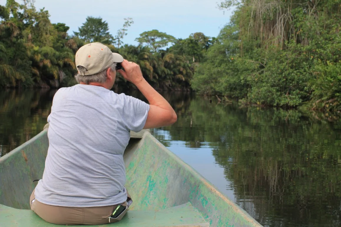 A person in a beige hat and gray shirt sitting in a green boat, looking through binoculars at the water and green trees on a river.