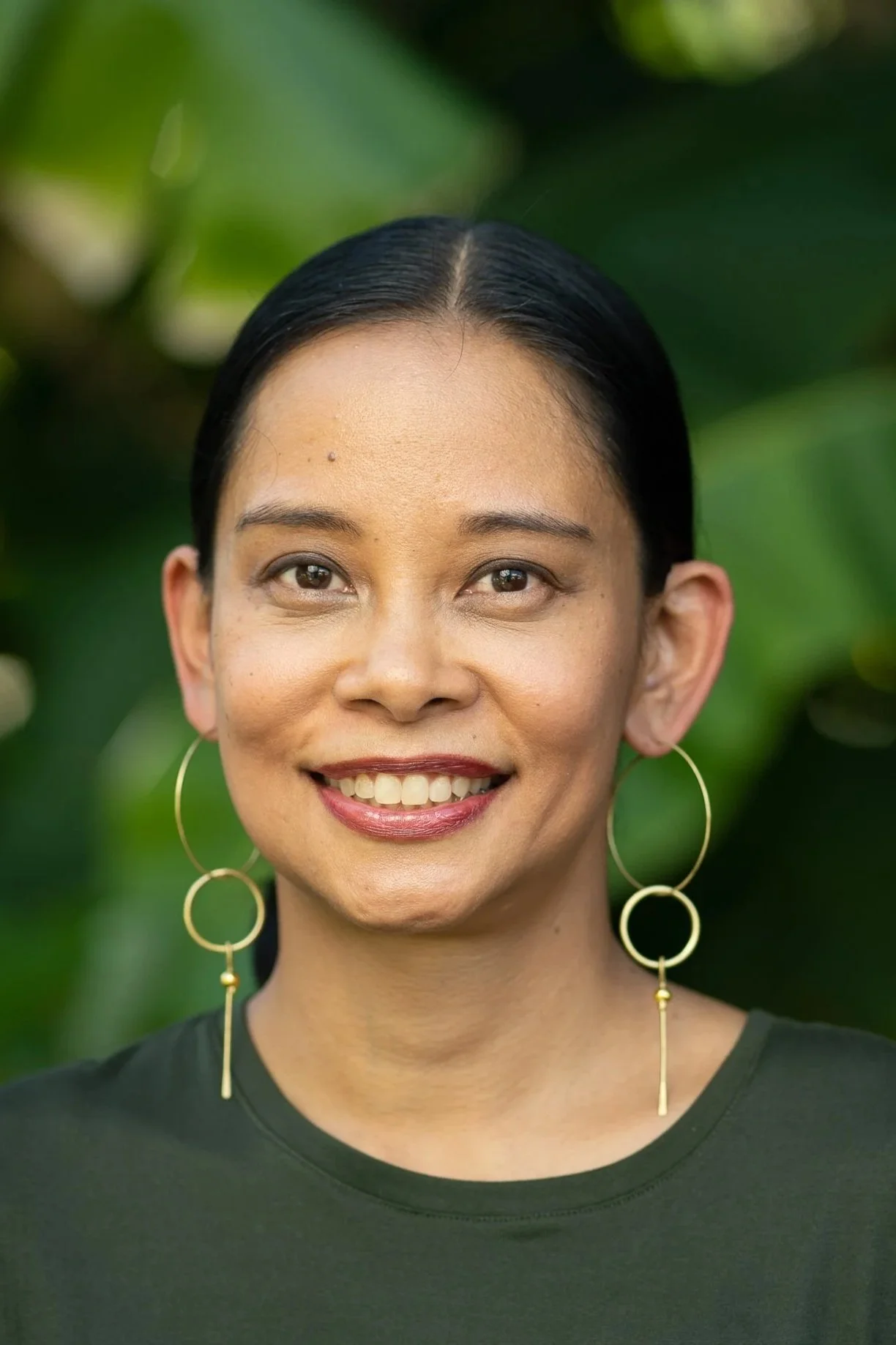 Close-up portrait of a woman smiling, wearing large hoop earrings and a dark t-shirt, with green foliage background.
