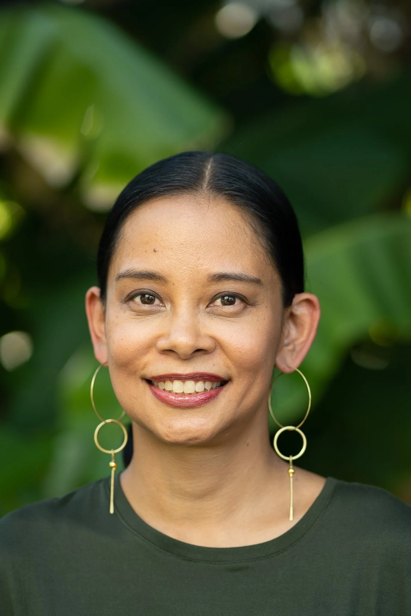 Close-up portrait of an Asian woman smiling, wearing large gold hoop earrings, with a blurred green leafy background.