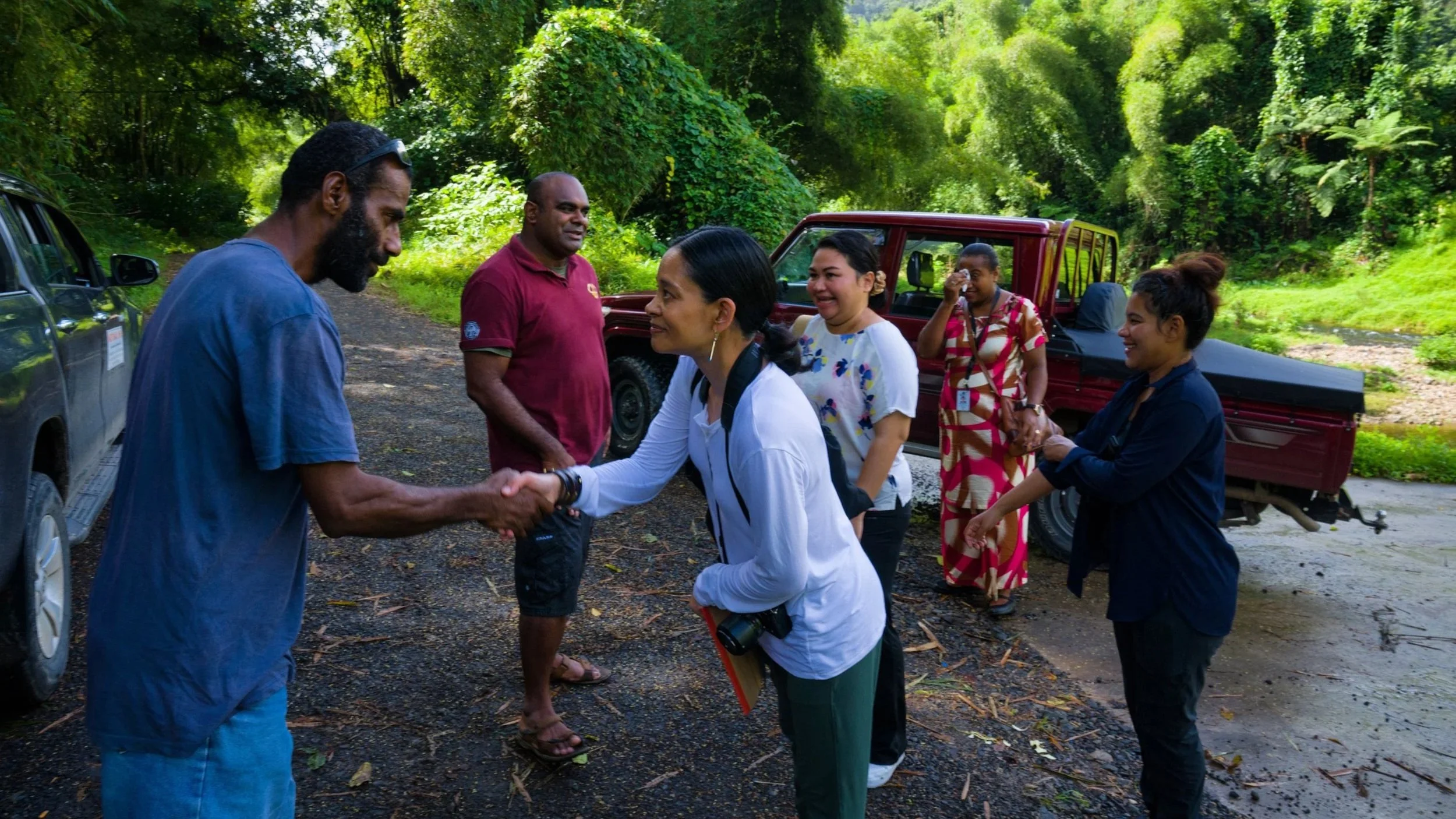 Two people shaking hands in an outdoor setting with a group of people and vehicles, surrounded by green foliage.