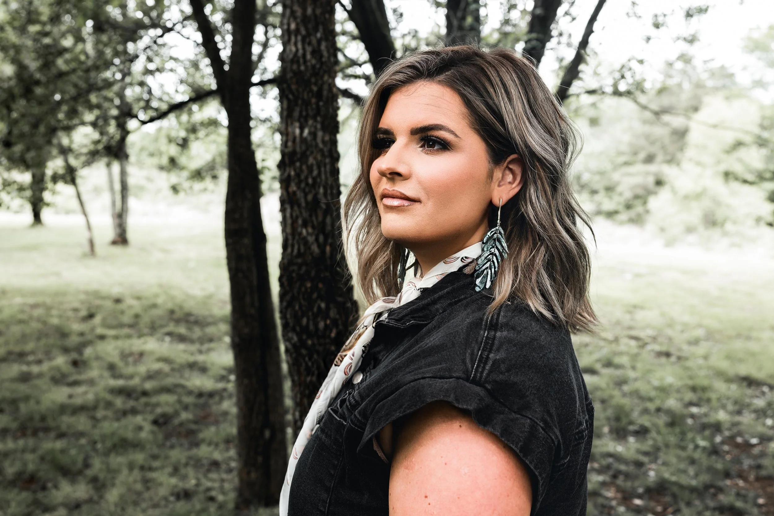 A woman with wavy light brown hair and earrings outdoors near trees, wearing a black denim jacket and a scarf with a feather pattern.