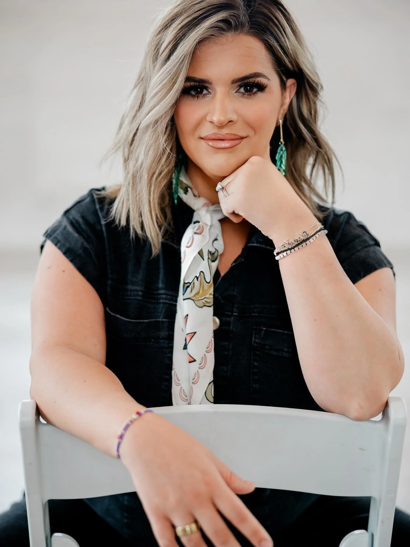A woman with blonde hair and makeup sitting at a table with her chin resting on her hand, wearing jewelry, earrings, ring, and a black top.