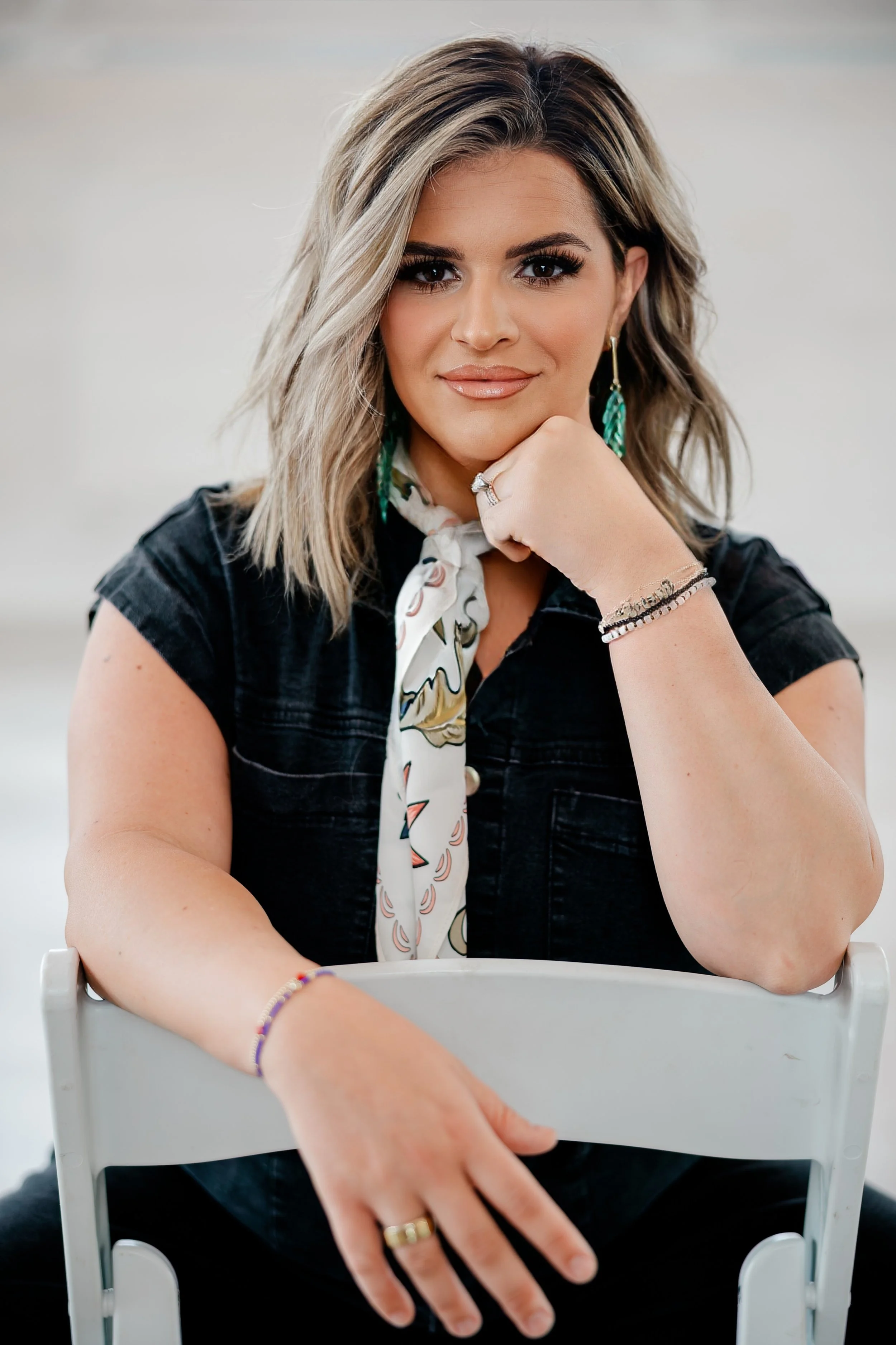 A woman with blonde, shoulder-length hair and olive skin is sitting at a table, resting her chin on her hand. She has dark eye makeup, nude lipstick, and large turquoise earrings. She is wearing a black shirt, a white scarf with a floral pattern, and multiple bracelets and rings. There is a white chair in front of her, and the background is blurred.