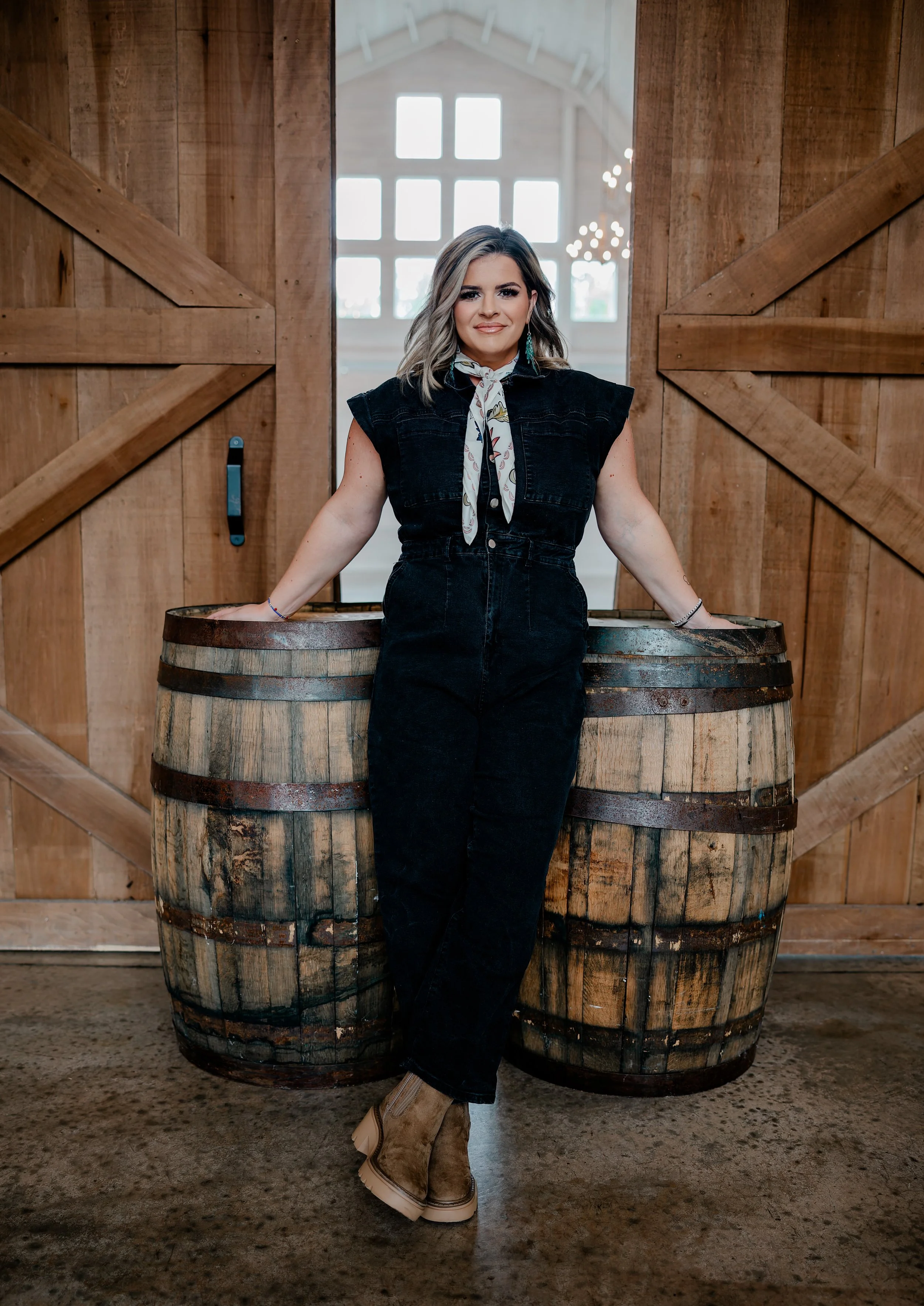 A woman standing in front of two wooden barrels inside a barn, with large wooden sliding doors behind her and a bright window in the background.