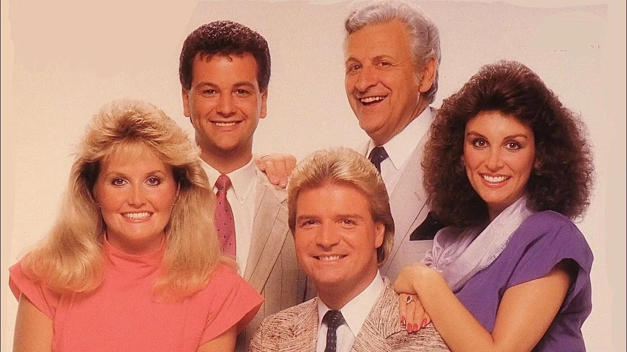 Six smiling adults posing together against a plain background, dressed in 1980s fashion. Three women and three men, with distinctive hairstyles including long, voluminous hair and a mustache on one man.