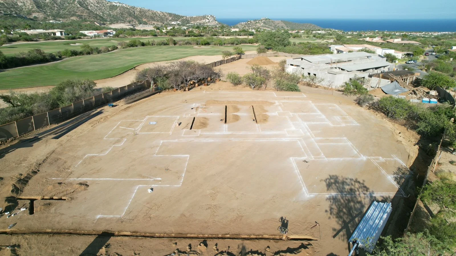 A construction site with outlines of building foundations marked with white chalk on the dirt ground, located near a golf course and residential neighborhood, with mountains and ocean in the background.