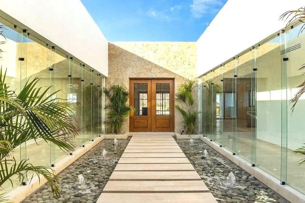 Modern indoor courtyard with a walkway across a shallow pond, glass walls on both sides, green plants, and a wooden double door at the end.
