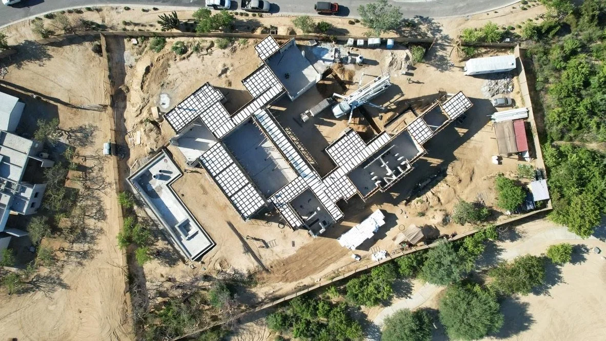 Aerial view of a house under construction with solar panels on the roof, surrounded by trees and dirt, with construction equipment and vehicles around.