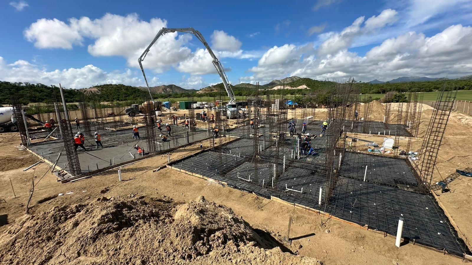 Construction site with workers pouring concrete for building foundations, with steel rebar structures in place and a concrete pump truck in the background under a partly cloudy sky.