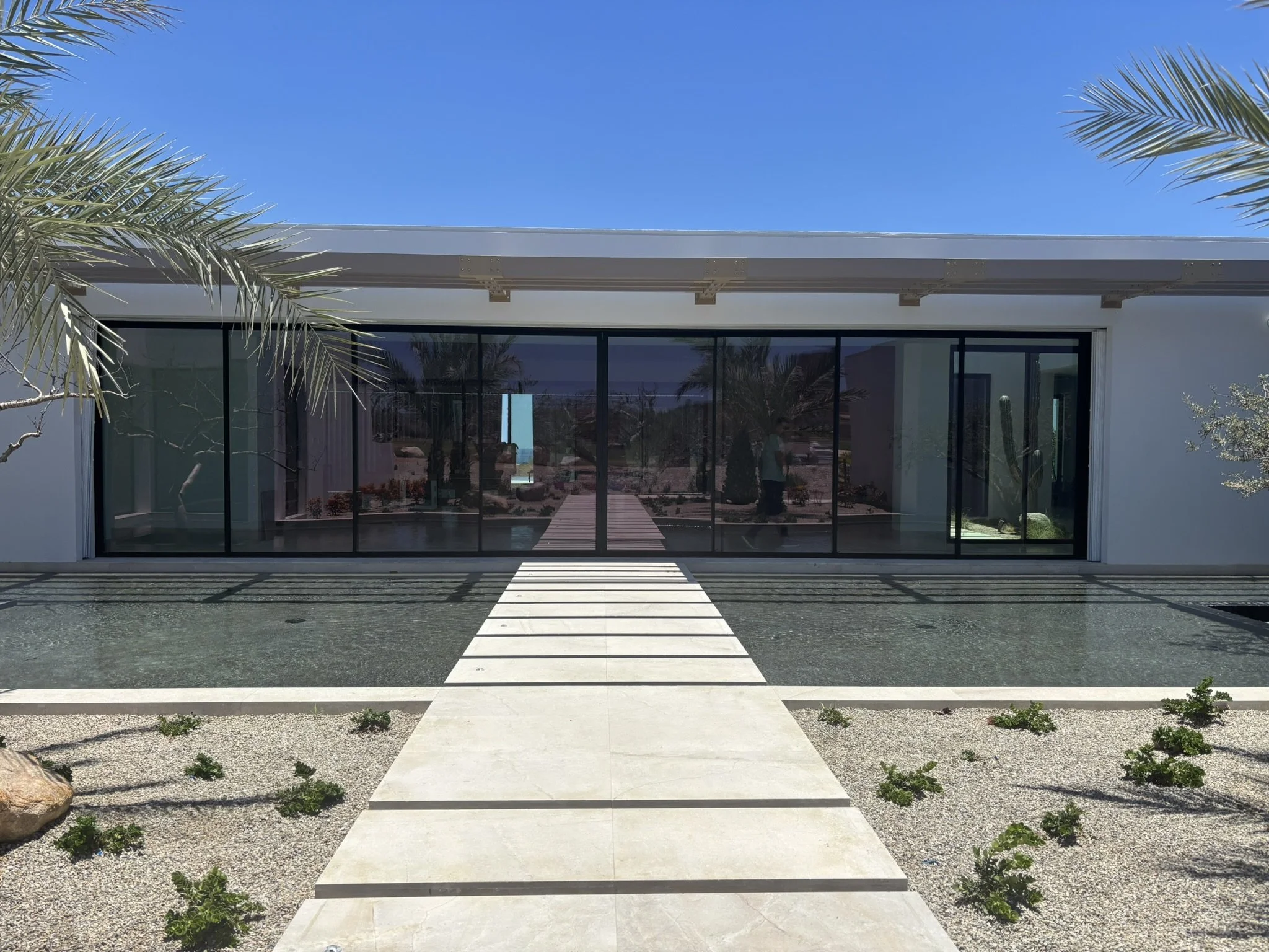 Modern house with large sliding glass doors, desert landscaping with small plants, palm trees, and a pathway leading to the entrance under a clear blue sky.