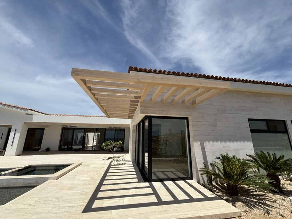 Modern house with a wooden pergola, large glass sliding doors, a pool, and desert plants under a partly cloudy sky.