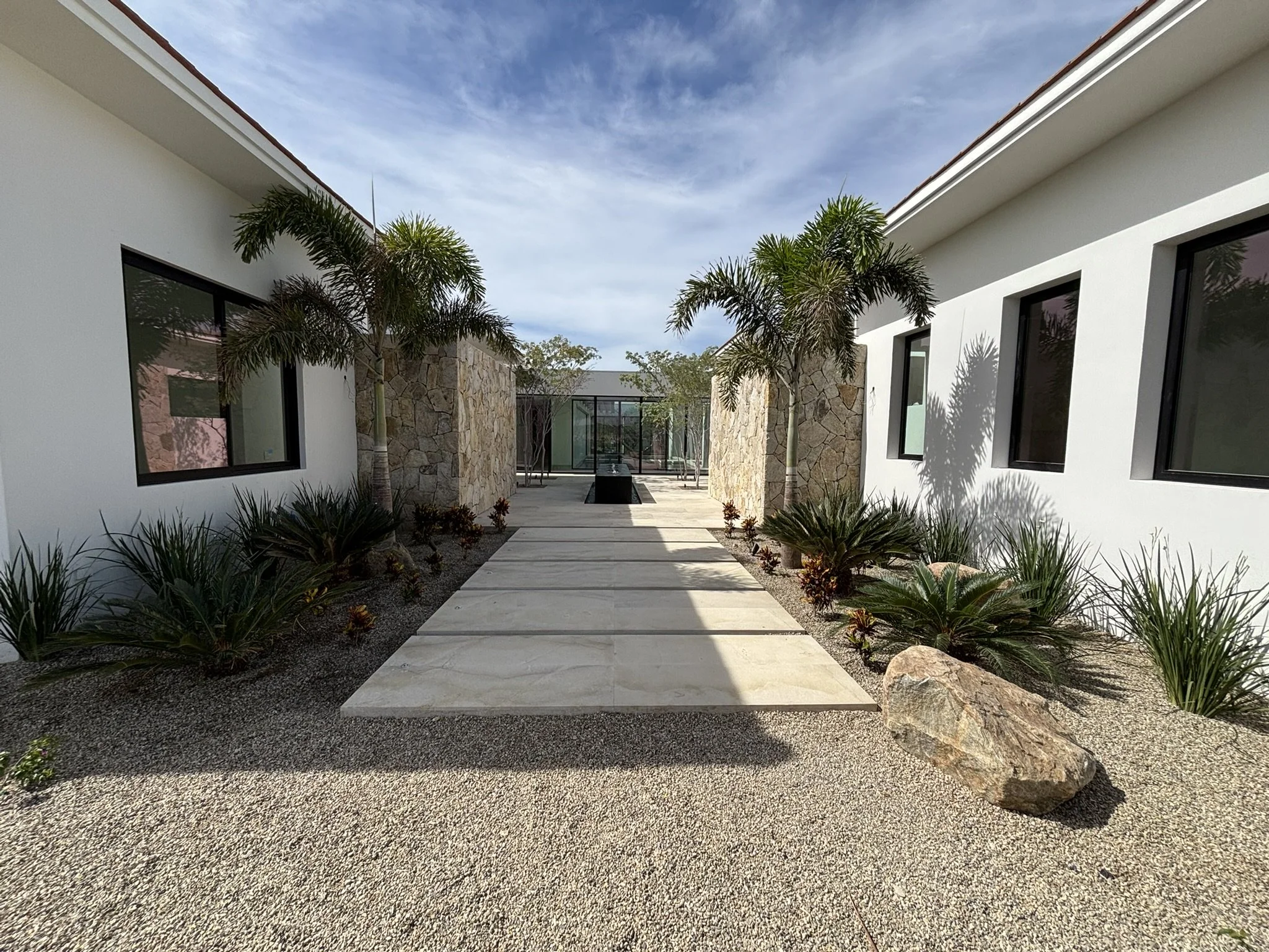Modern courtyard with a concrete walkway, white walls, and tropical plants, including palm trees, under a partly cloudy sky.